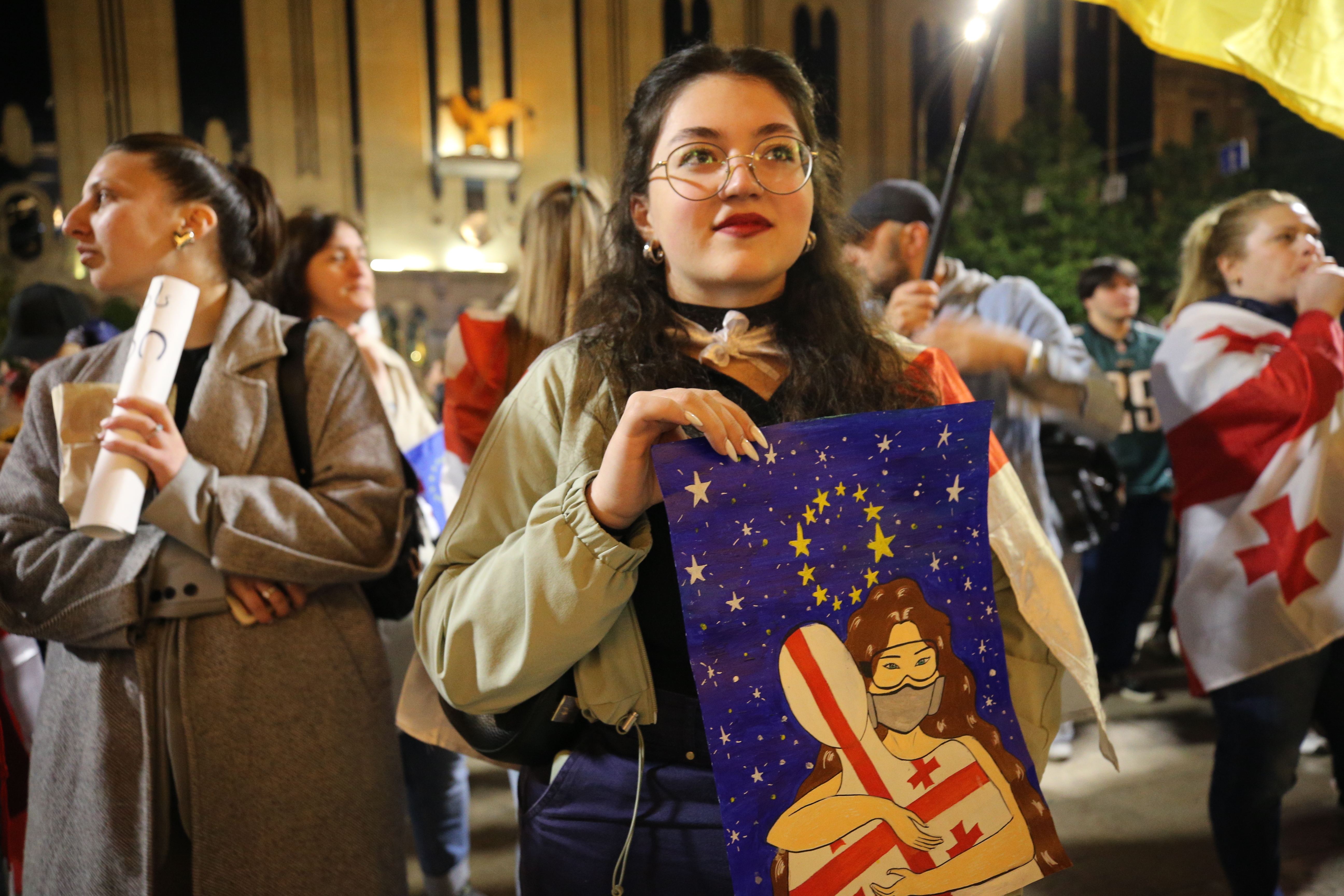 Eine junge Frau steht in mitten anderer Demonstrierenden und hält ein Plakat hoch, auf dem zwei sich umarmende Figuren, eine davon mit georgischer Flagge, vor blauem Nachthimmel und EU-Sternen zu sehen sind.