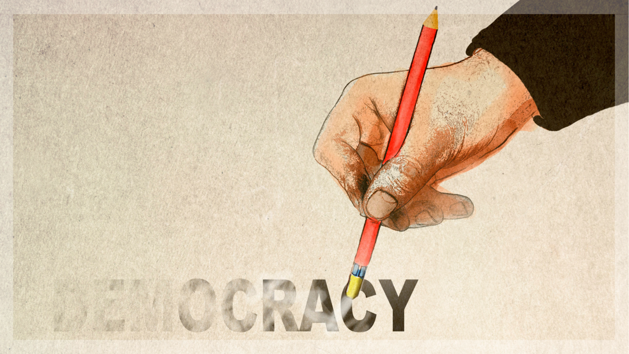 One hand holds a pencil and erases the word democracy.