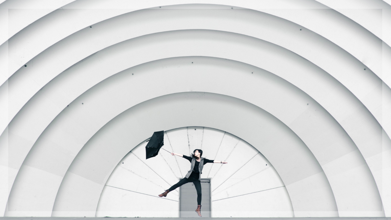 Frau mit Regenschirm springt vor einer großen grauen Wand, getragen vom starken Wind.